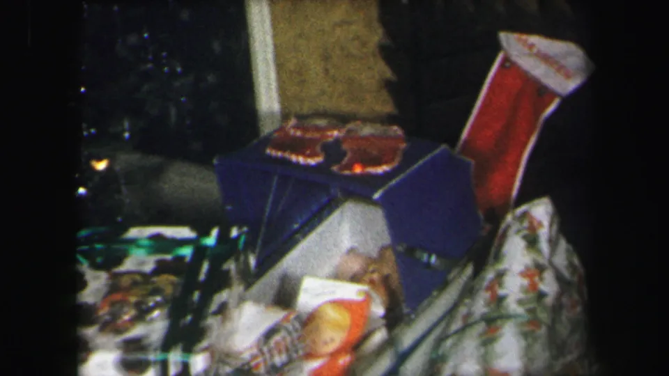 1971 Lynbrook home-movie frame showing a red Christmas stocking, gift boxes, patterned fabric, and dark living-room furniture.