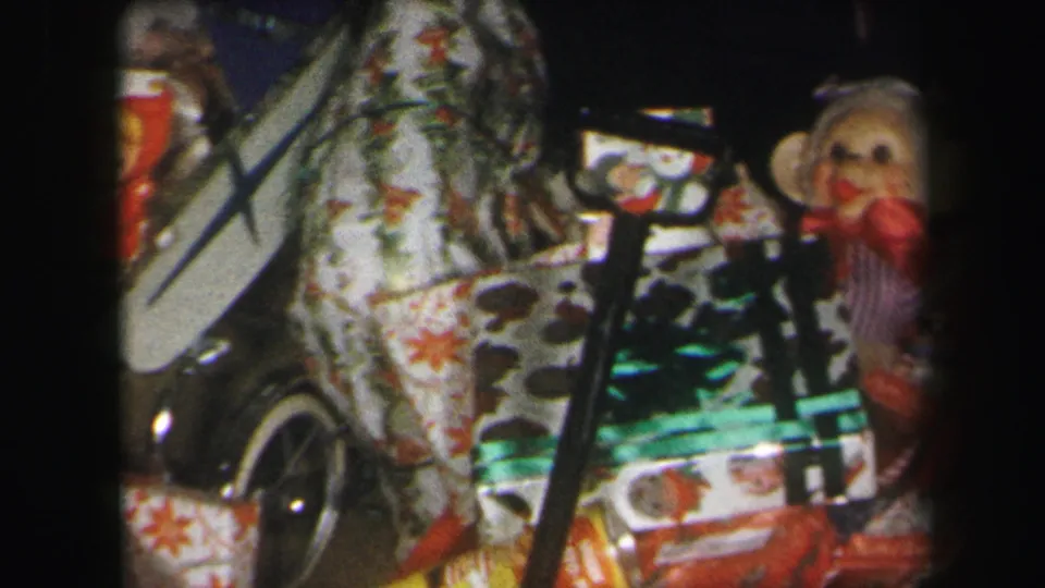 1971 Lynbrook home-movie frame showing a dense pile of Christmas presents, patterned fabric, wagon parts, and a sock monkey at right.