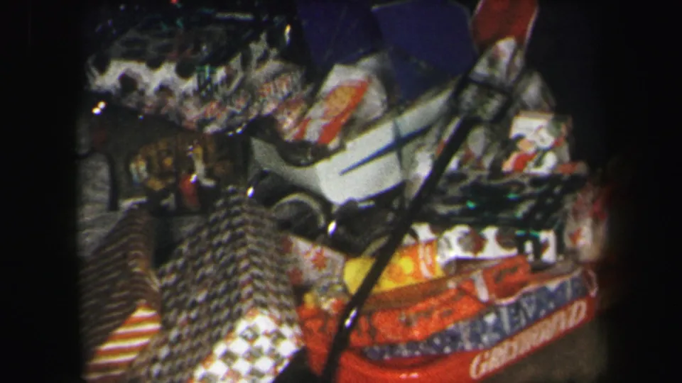 1971 Lynbrook home-movie frame with red and patterned Christmas presents, cards, a small doll, and a white toy stroller in soft focus.