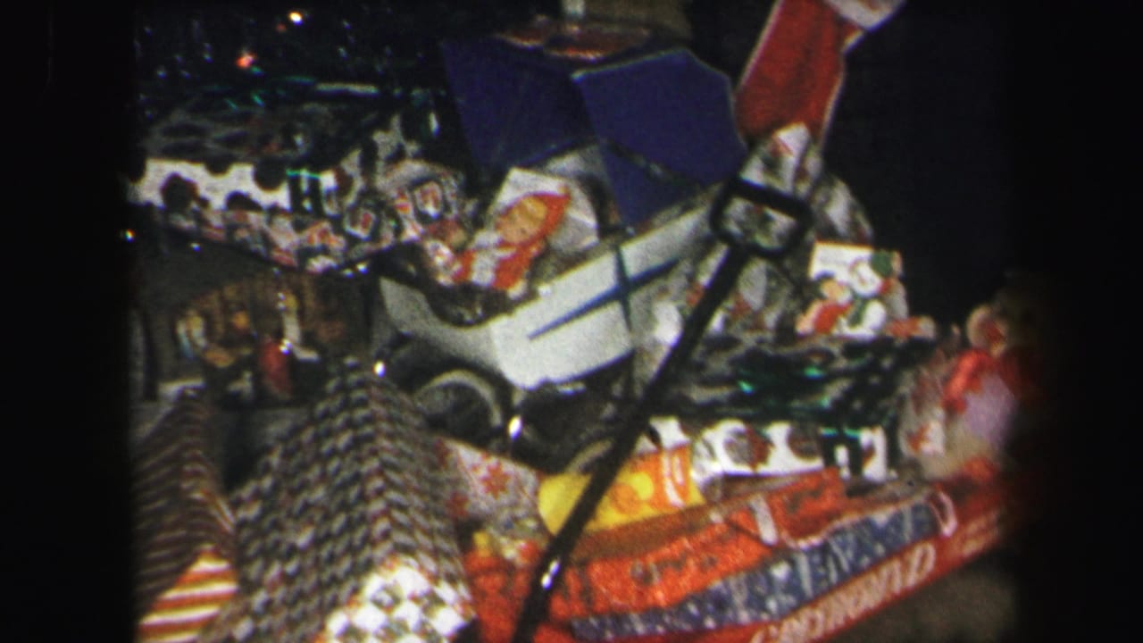 1971 Lynbrook home-movie frame showing a red wagon filled with wrapped gifts, toys, and a white toy stroller in a dim interior.