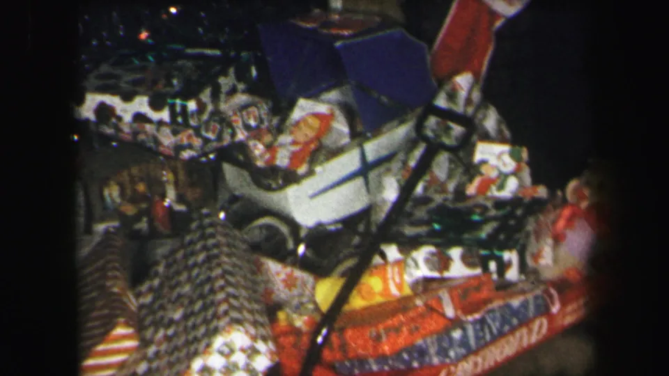 1971 Lynbrook home-movie frame showing a red wagon filled with wrapped gifts, toys, and a white toy stroller in a dim interior.