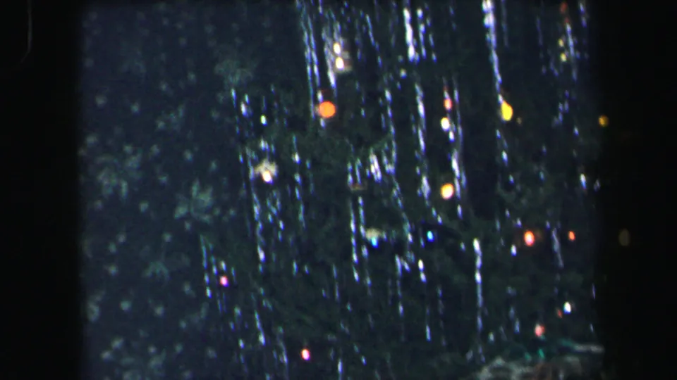1971 Lynbrook home-movie frame showing a tinsel-covered Christmas tree with small colored lights against a dark interior.