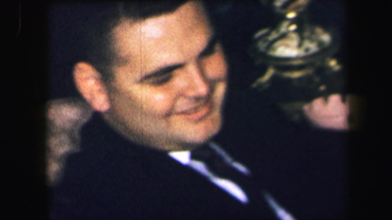 In 1971 Lynbrook home-movie footage, an adult man in a dark suit smiles while seated in a patterned chair indoors.