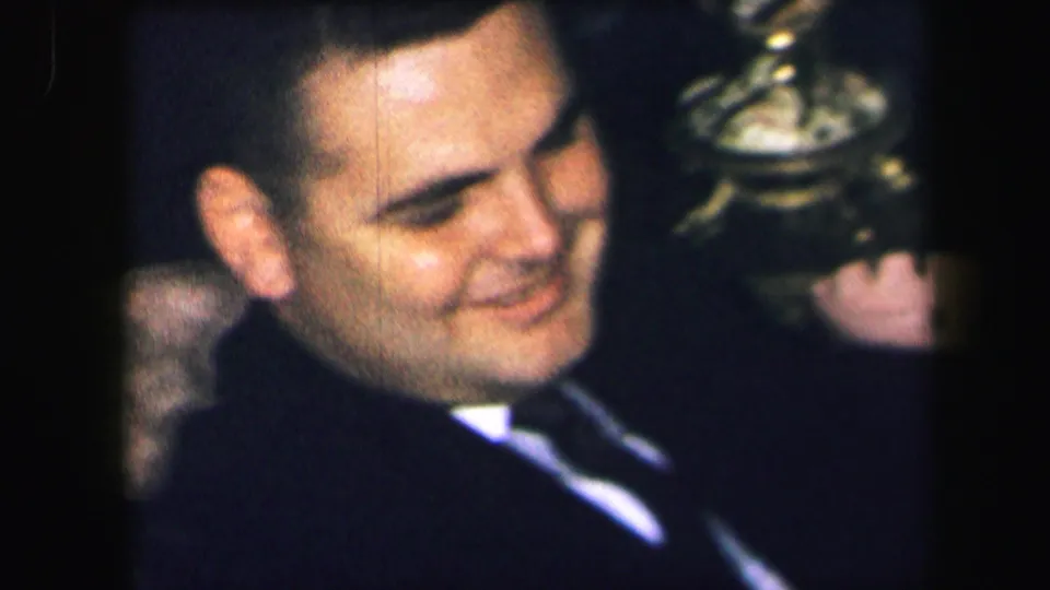 In 1971 Lynbrook home-movie footage, an adult man in a dark suit smiles while seated in a patterned chair indoors.