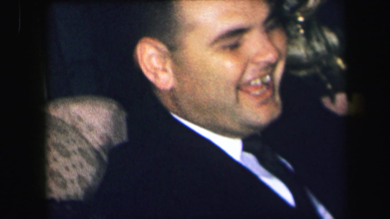 The seated man laughs in a dark suit and white shirt during the 1971 Lynbrook indoor home-movie scene.