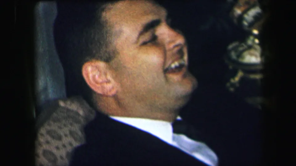 An adult man in Lynbrook, New York, leans back laughing in a chair in grainy 1971 home-movie footage.