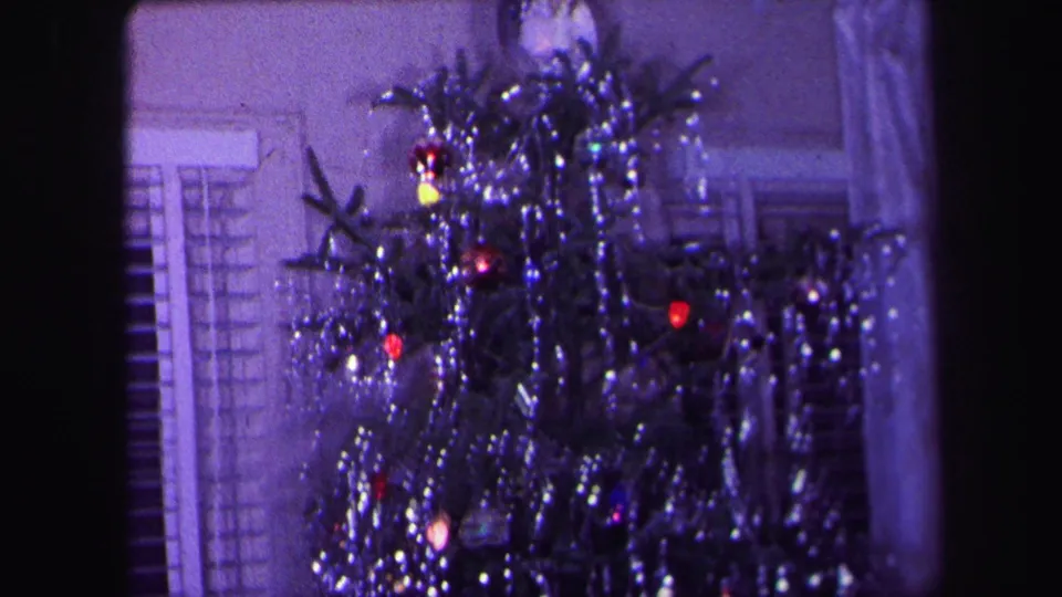 1971 Lynbrook home-movie frame showing a decorated indoor Christmas tree with tinsel, colored lights, red ornaments, and blinds in the background.