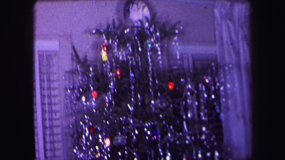 1971 Lynbrook home-movie frame showing a tinsel-covered Christmas tree with colored lights near curtains and a window.
