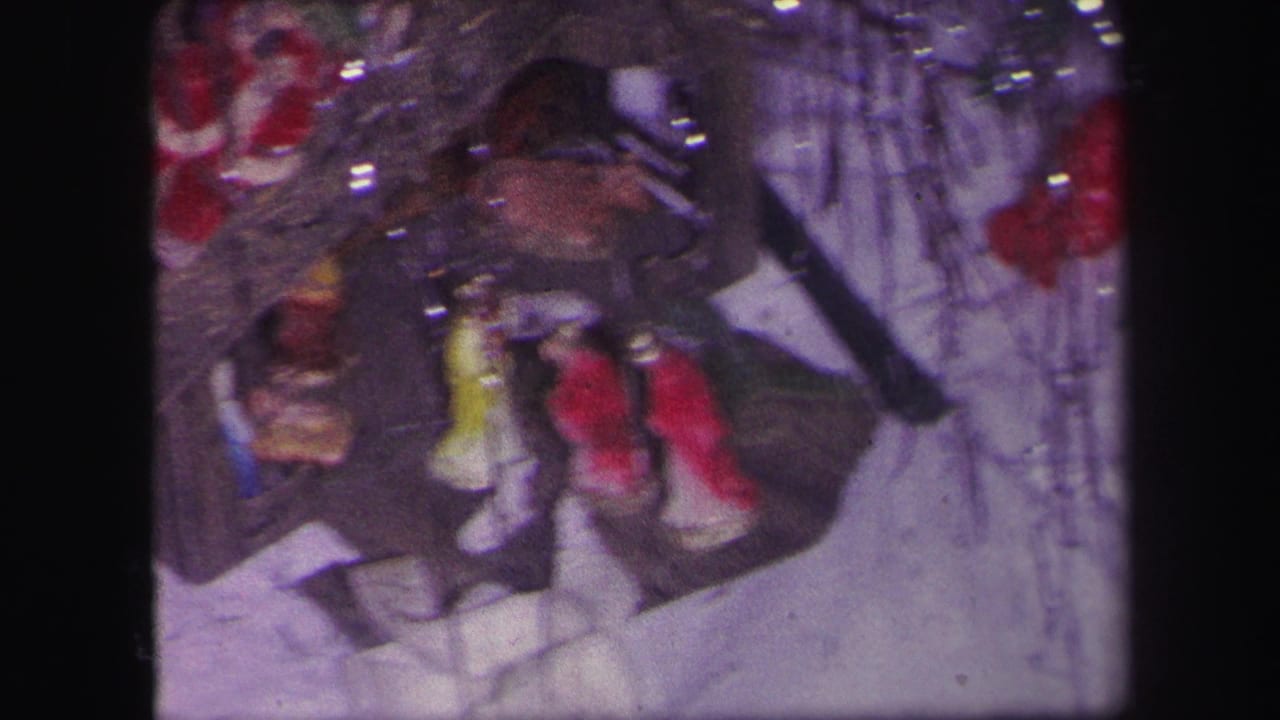 1971 Lynbrook home-movie frame showing a toy train setup with small red and yellow figures near tinsel and a white tree skirt.