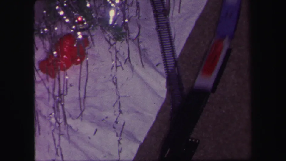 1971 Lynbrook frame with tinsel, red decorations or toy parts, a train track, and white cloth seen in soft purple-tinted 8mm color.
