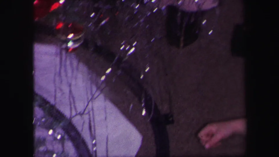 Handheld 8mm frame showing the toy train area, tinsel, track, white tree skirt, and a person's hand at the edge of the scene.