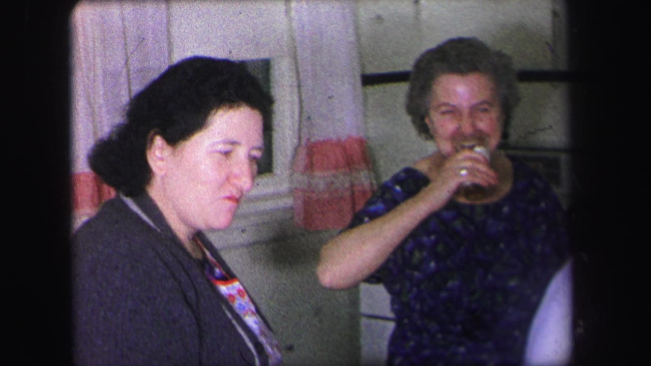 Two women seated indoors, one looking down and one smiling while drinking from a cup.
