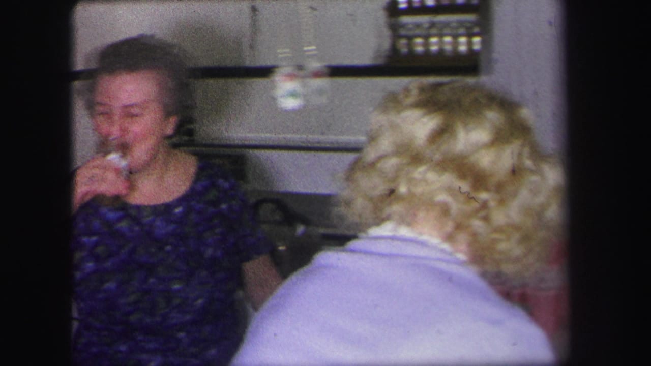 A woman drinks from a cup while another person with blond hair faces away in the foreground.