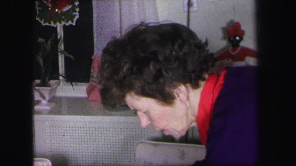 Inside the 1971 Lynbrook home, a woman leans forward near a window decorated with a red wreath and a small red-dressed doll.
