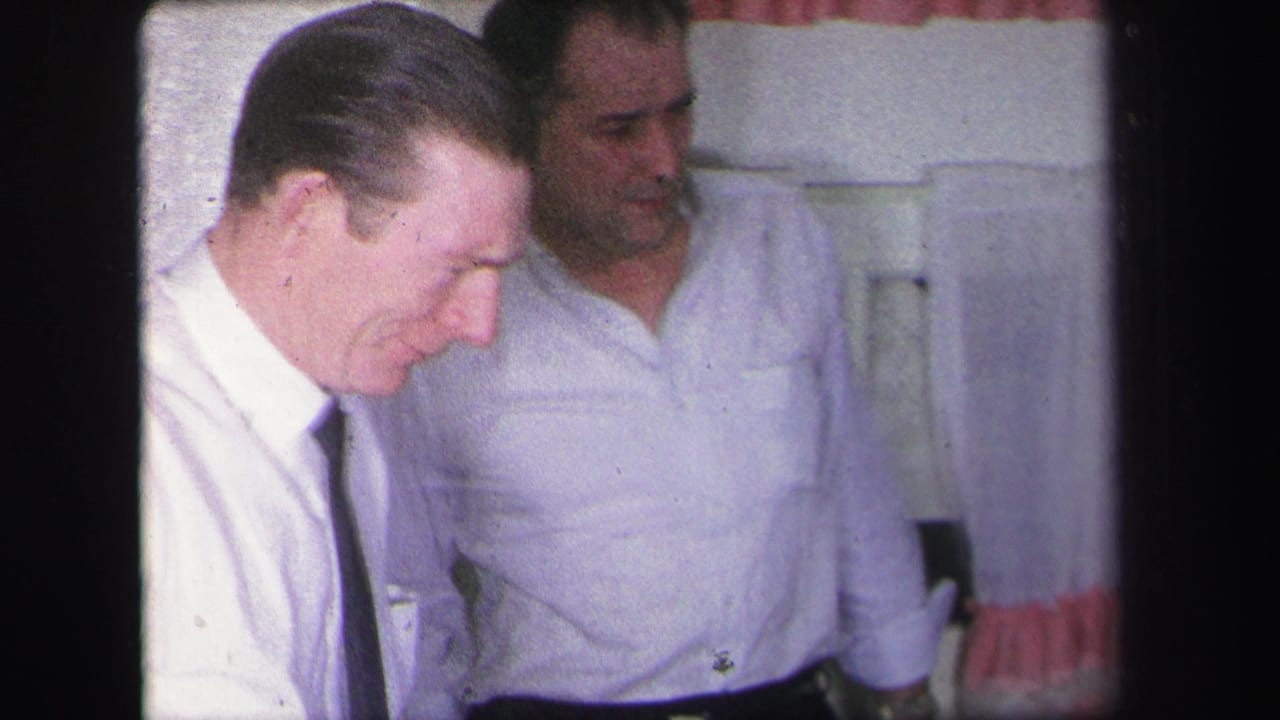 In a Lynbrook, New York home-movie frame from 1971, two adult men in white shirts stand close together in a domestic interior.