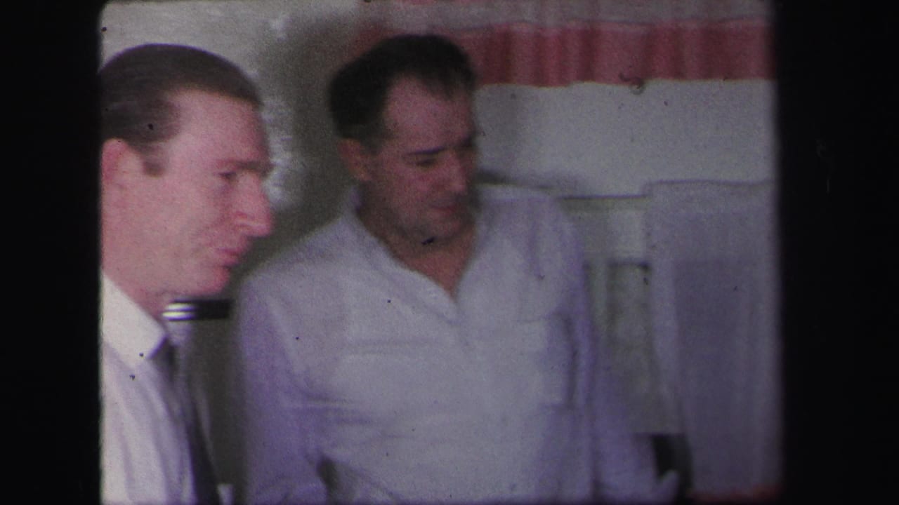 Two men in white shirts stand indoors, with one turned partly toward the camera.