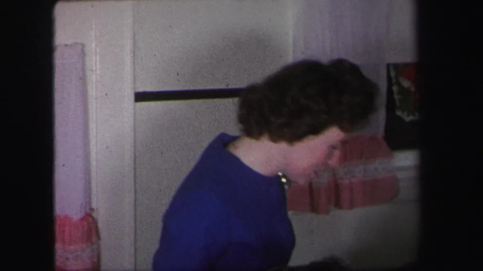 A woman in a blue top leans forward in a Lynbrook domestic interior, captured in soft 1971 8mm home-movie color.