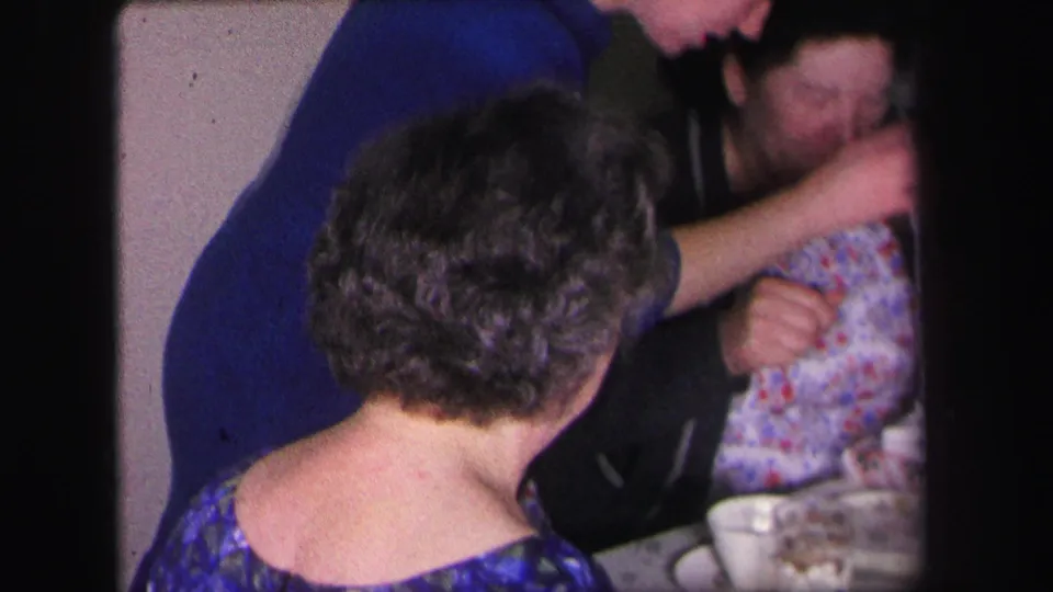 Several adults sit or lean around a table in a 1971 Lynbrook home interior, with cups and close handheld framing visible.