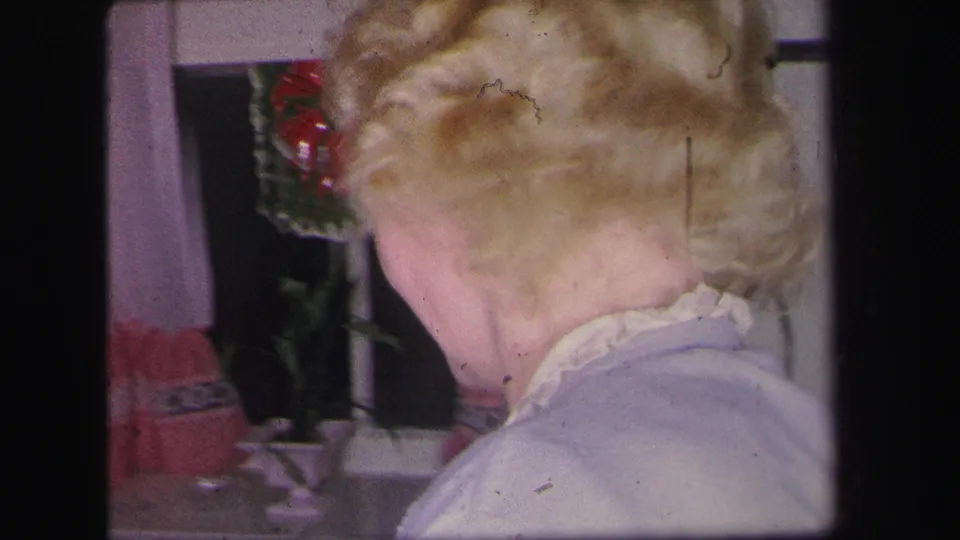The camera frames the back of a blonde woman near pink-trim curtains and a wreath in the private Lynbrook home interior.