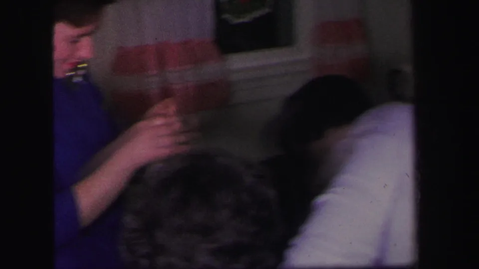 A woman in blue holds up or handles a necklace as seated women gather nearby in the 1971 Lynbrook home-movie scene.