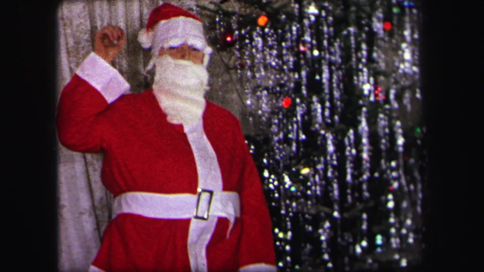 In a 1971 Lynbrook home movie, a child in a red Santa suit and white beard waves beside a decorated tinsel Christmas tree.