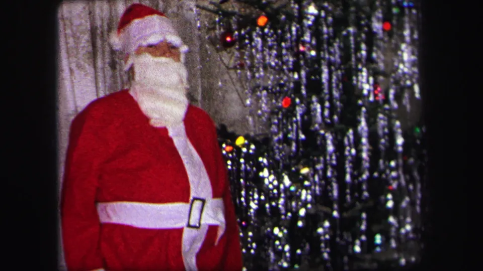 A child in a red Santa costume stands near curtains and a lit tinsel Christmas tree in 1971 Lynbrook family footage.