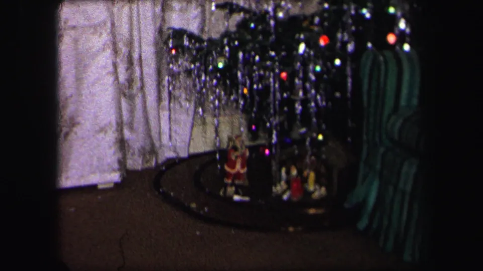 The base of the Christmas tree shows small holiday figurines, a toy train track, curtains, and a green striped object in a private home.