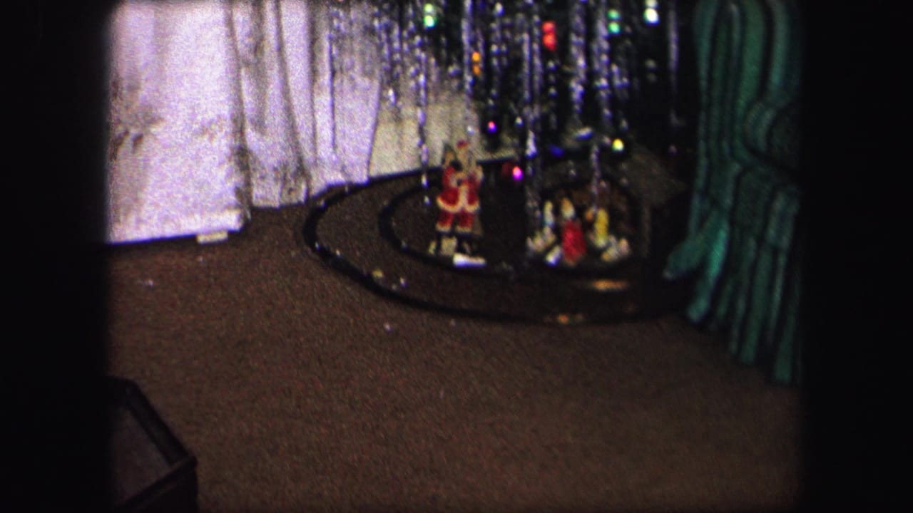 1971 Lynbrook home-movie frame showing the base of a Christmas tree, small holiday figures, and a train track on a carpeted floor.