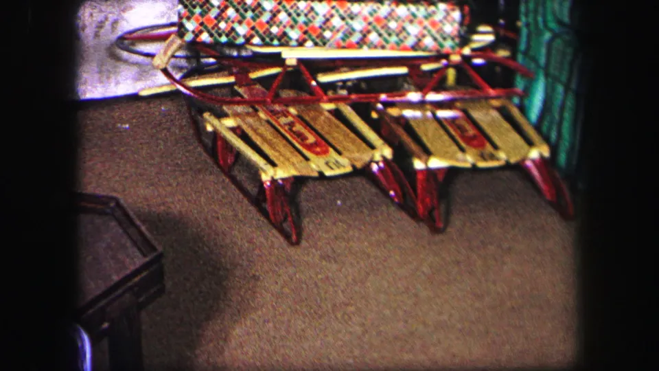 1971 Lynbrook interior frame with a colorful plaid blanket on one sled and a second wooden sled nearby on carpet.