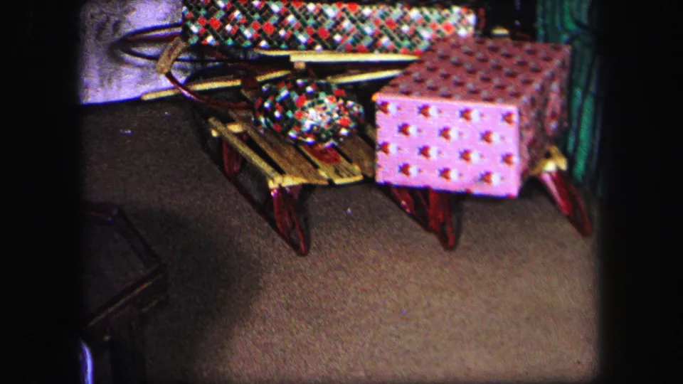 1971 Lynbrook home-movie frame with a large red-and-white wrapped box beside wooden sleds and a plaid blanket.