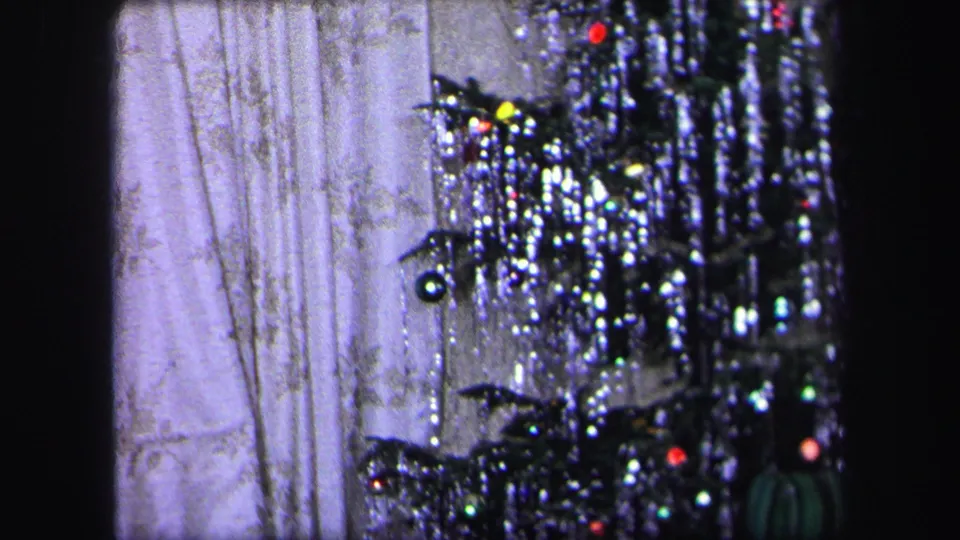 1971 Lynbrook home-movie frame showing a tinsel-covered Christmas tree with small ornaments against patterned curtains.
