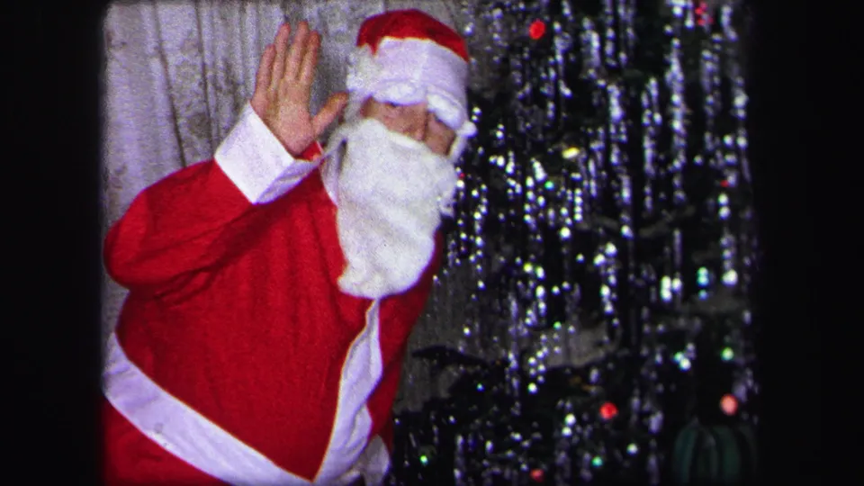 1971 Lynbrook home-movie frame showing a person in a Santa suit waving near a tinsel Christmas tree in a private living room.