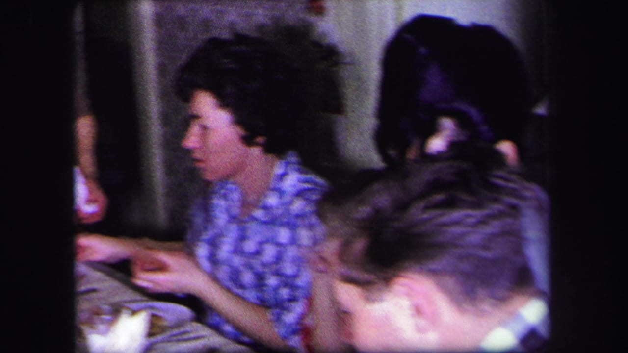 1971 Lynbrook home-movie frame showing adults at a dinner table, including a woman in a blue patterned blouse and tableware in front of her.
