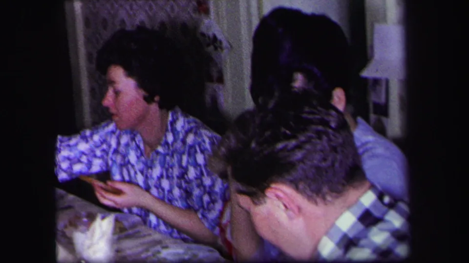 1971 Lynbrook dining-room frame with adults eating and conversing at a crowded table inside a private home.