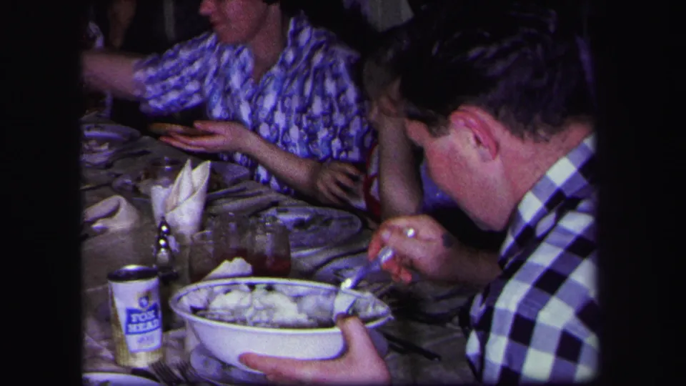 Close home-movie view of a man eating at a 1971 Lynbrook family dinner table with dishes, glasses, and napkins visible.