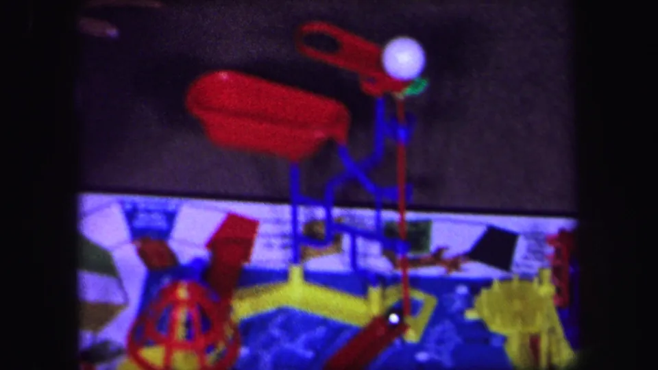 1971 Lynbrook, New York 8mm frame showing the lower portion of a colorful mechanical board game with red levers, yellow parts, and blue structure.