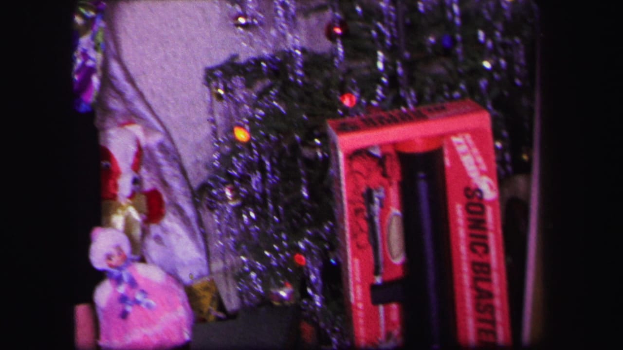 1971 Lynbrook home-movie frame with a woman near a small Christmas tree, a pink doll, tinsel, and a red toy box.