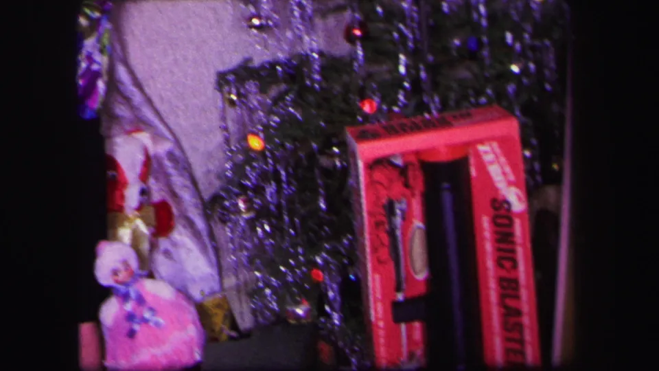 1971 Lynbrook home-movie frame with a woman near a small Christmas tree, a pink doll, tinsel, and a red toy box.