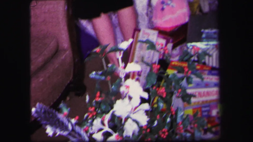 1971 Lynbrook home-movie frame of a woman arranging toys and gifts beside a small decorated Christmas tree.