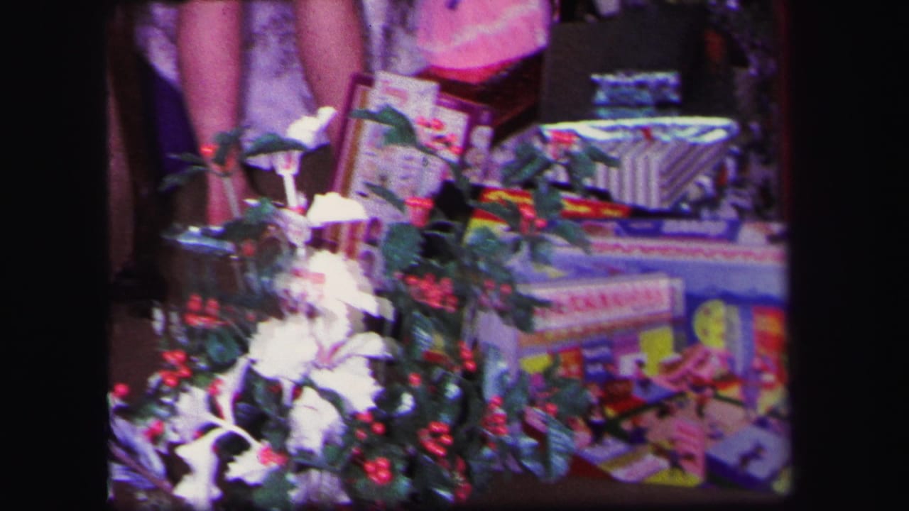 1971 Lynbrook home-movie frame showing Christmas presents, holly, tinsel, and toy packaging with partial legs visible behind the display.