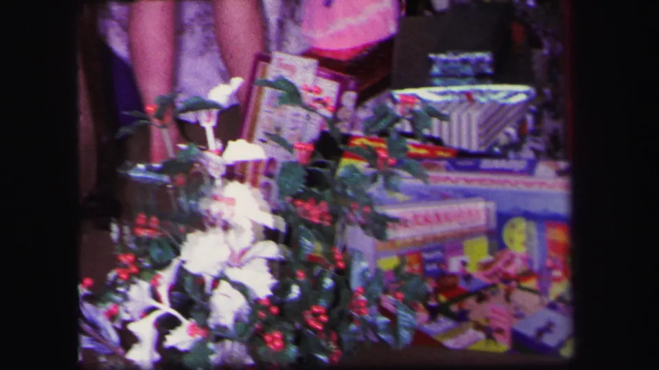 1971 Lynbrook home-movie frame showing Christmas presents, holly, tinsel, and toy packaging with partial legs visible behind the display.