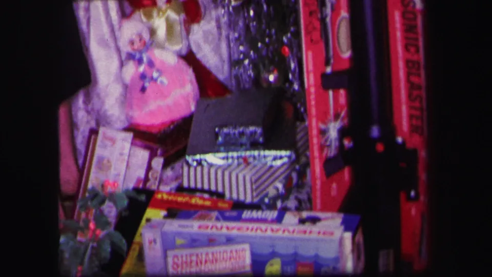 1971 Lynbrook home-movie frame showing the Christmas toy display with a pink doll, Shenanigans box, holly, tinsel, and red toy blaster.