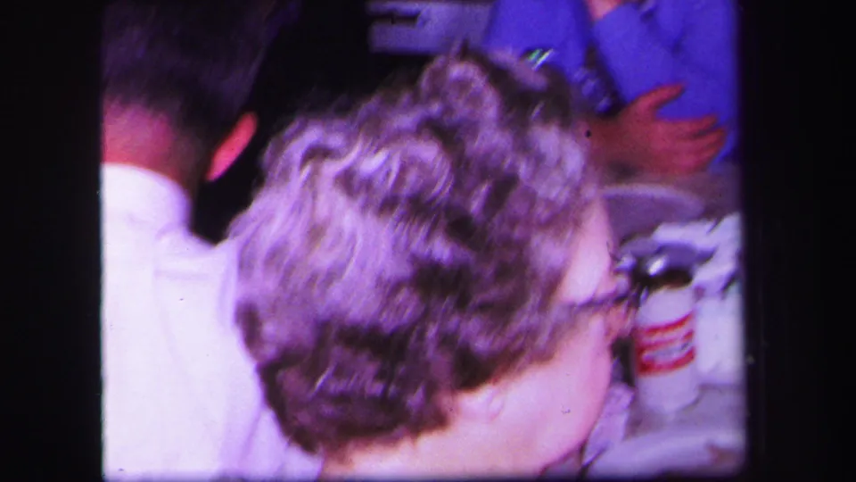 1971 Lynbrook home-movie frame of adults seated closely near a table during a family party.