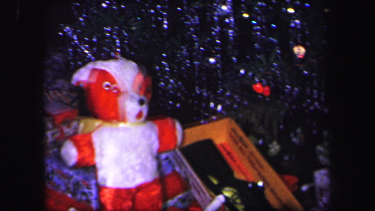 1971 Lynbrook home-movie frame showing a red and white stuffed dog toy beneath a tinsel Christmas tree with gifts nearby.