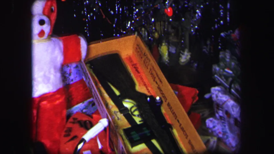 1971 home-movie frame with a boxed toy BB gun and partial stuffed dog figure under a tinsel Christmas tree in Lynbrook, New York.