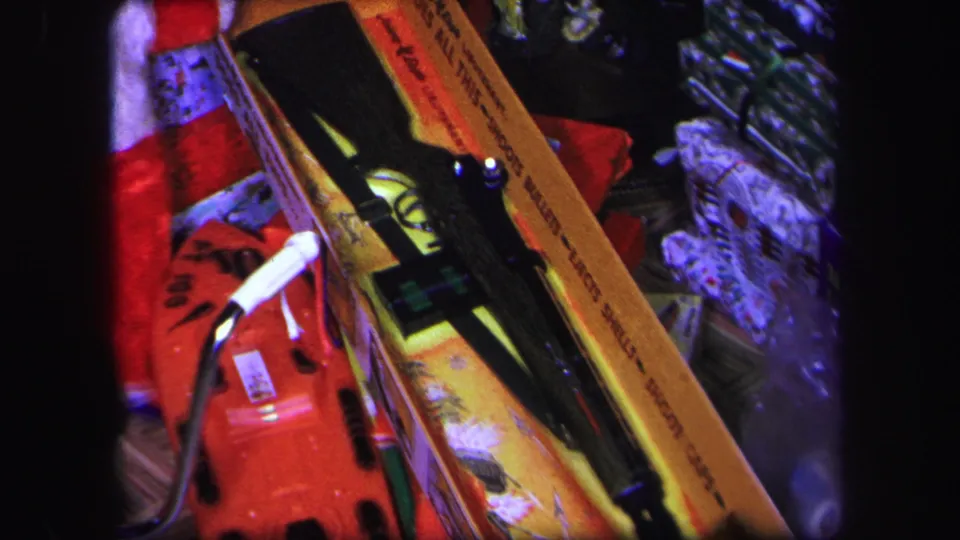 Final 8mm frame focuses on the boxed toy BB gun beneath the Lynbrook Christmas tree, with tinsel, lights, and holiday ornaments around it.