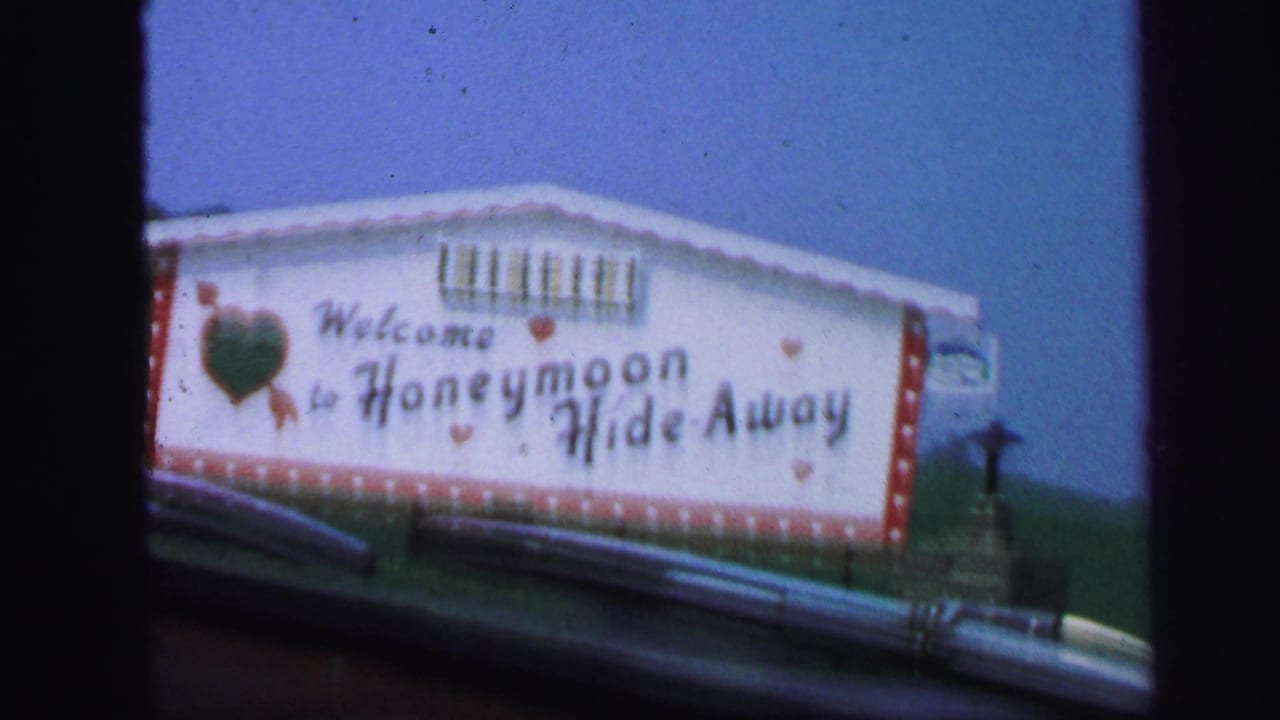 White roadside sign reading Welcome to Honeymoon Hide-Away beside a road.