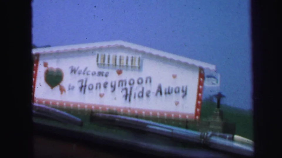 1971 8mm passenger view of a large white Honeymoon Hide-Away sign with red trim, a green heart, blue sky, and roadside guardrail.