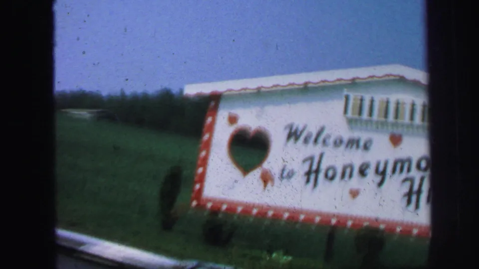 Soft 1971 8mm frame showing a close passenger-side view of the Honeymoon Hide-Away sign, with green grass, blue sky, and motion blur.