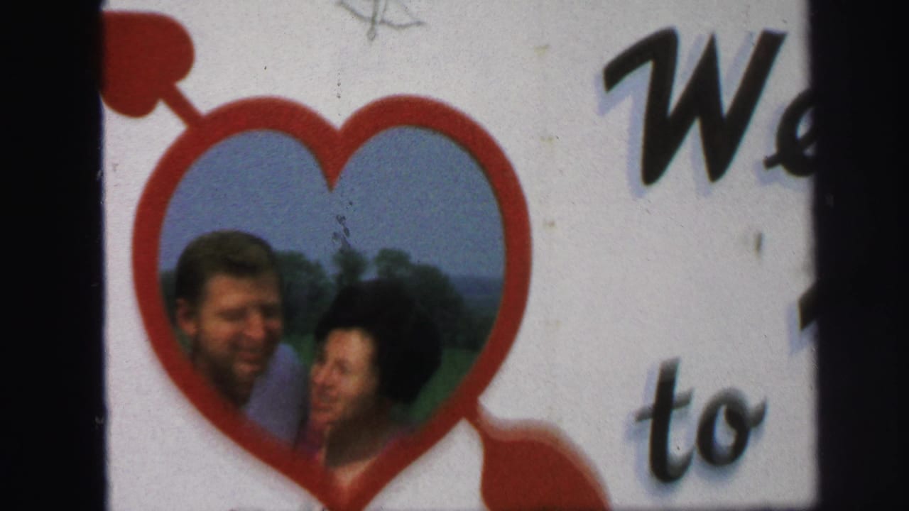 1971 Lynbrook home-movie frame showing a man and woman posing inside a red heart-shaped cutout with grass and sky behind them.